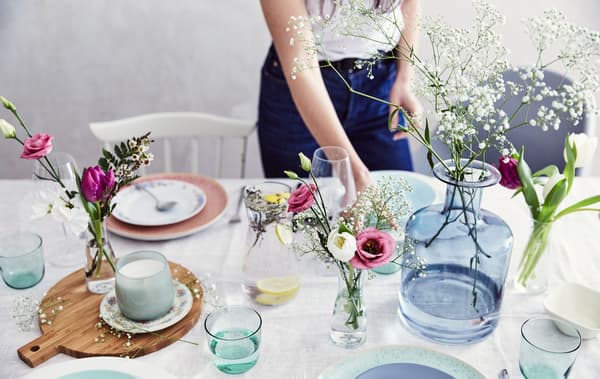A table set with pastel tableware and fresh flowers in glass vases of different shapes and sizes, with a wooden board and cotton tablecloth.