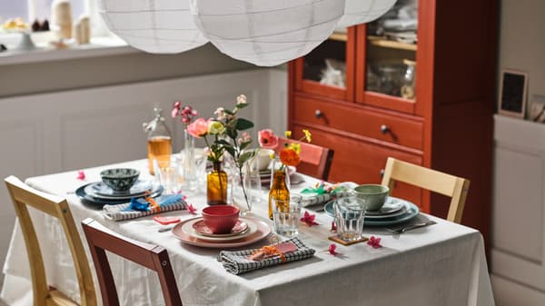 A table with a natural/beige tablecloth set with mixed dinnerware in different colours, POKAL glasses and artificial flowers.