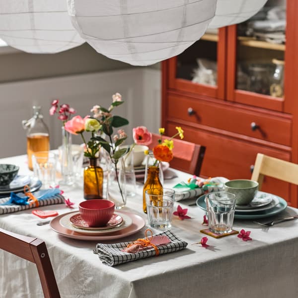 A table with a natural/beige tablecloth set with mixed dinnerware in different colours, POKAL glasses and artificial flowers.