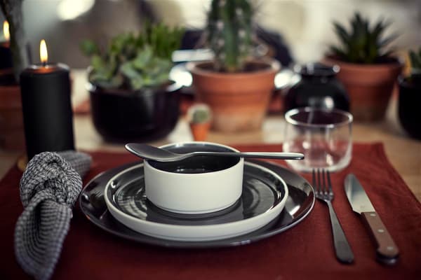 A tablesetting with a black plate, a dark chocolate desert plate and a small black bowl with a spoon on top, layed out on a red tablemat.