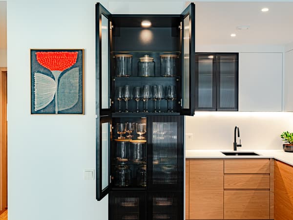 A tall kitchen cabinet with anthracite reeded glass doors, open to reveal glassware lit by integrated lighting, set against light walls beside the kitchen area.