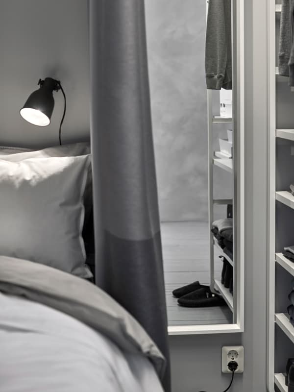 A tall, narrow mirror placed between bed and open wardrobe. Black spotlight over bed and grey curtain beside it.