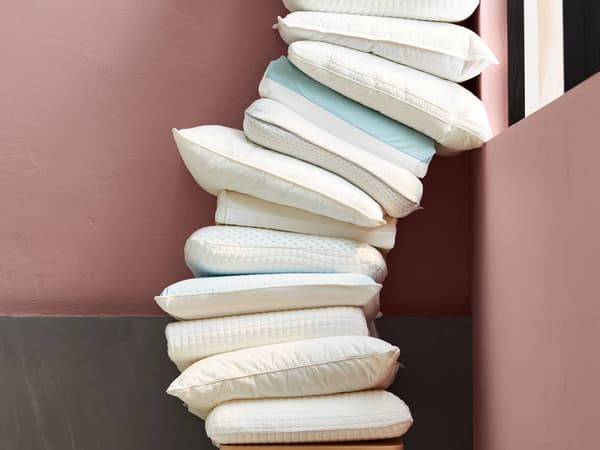 A tall stack of white and light blue pillows leans against a pink and gray wall, creating a cozy arrangement.