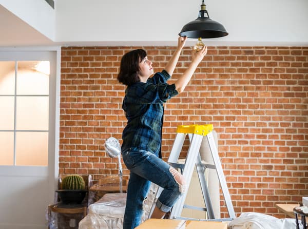 A Task Rabbit tasker changing a light bulb of a ceiling lamp shade while standing on a ladder, in a house that is being renovated.