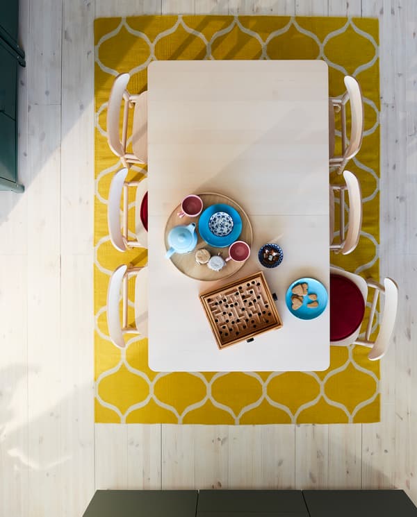 A teapot, cups, biscuits and a board game on a table in birch, six chairs in birch, two red chair pads and a yellow rug.