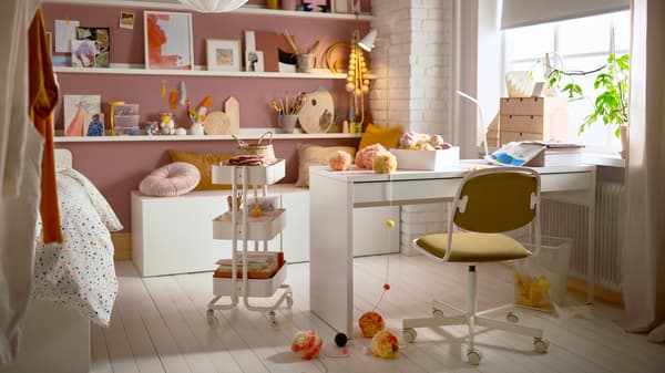 A teenager’s room with a desk, a trolley, picture ledges with artsy decorations, cushions, and a swivel chair.