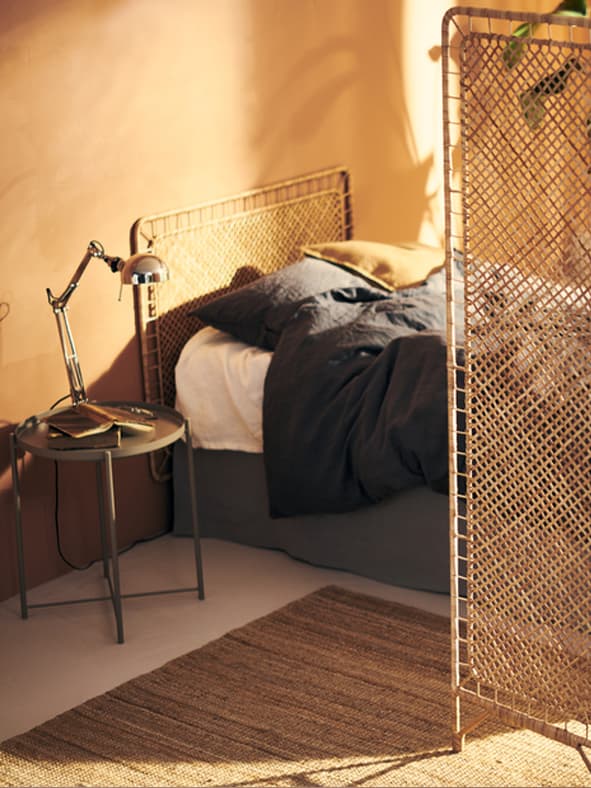A TERTIAL work lamp is placed on the bedside table. The super single bed with a TOLKNING headboard is partially concealed by the TOLKNING Room divider, which is also made of rattan.