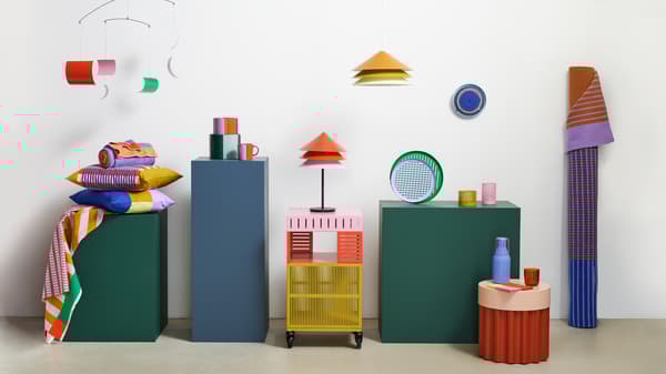 A TESAMMANS table lamp is on top of a storage unit on castors beside other colourful TESAMMANS items placed on pedestals.