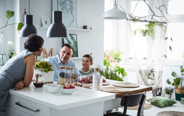 A three-person family around the kitchen island in a large, bright, white kitchen with lots of plants and green accents.