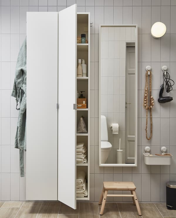 A tiled wall with LILLÅNGEN high cabinets with side-hooks, a white mirror, a round wall lamp and TISKEN hooks and basket.