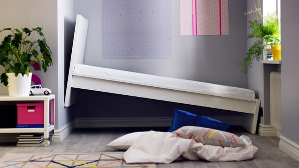 A tilted white bed leans against a gray wall in a modern room with geometric artwork. A plant, colorful box, and scattered pillows create a quirky, playful vibe.