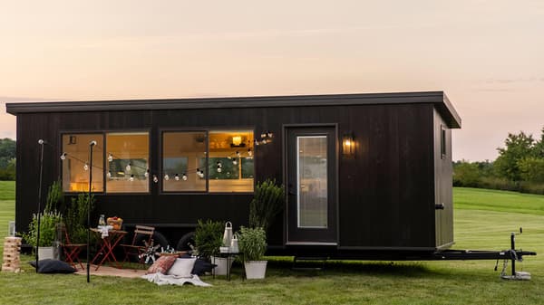 A tiny home with string lights and red outdoor furniture in front, parked on grass with a colorful sky above.