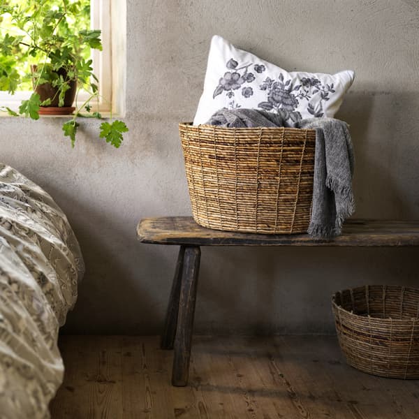 A TJILLEVIPS basket made from banana fibre is filled with blankets and a white floral cushion and placed on a bench.