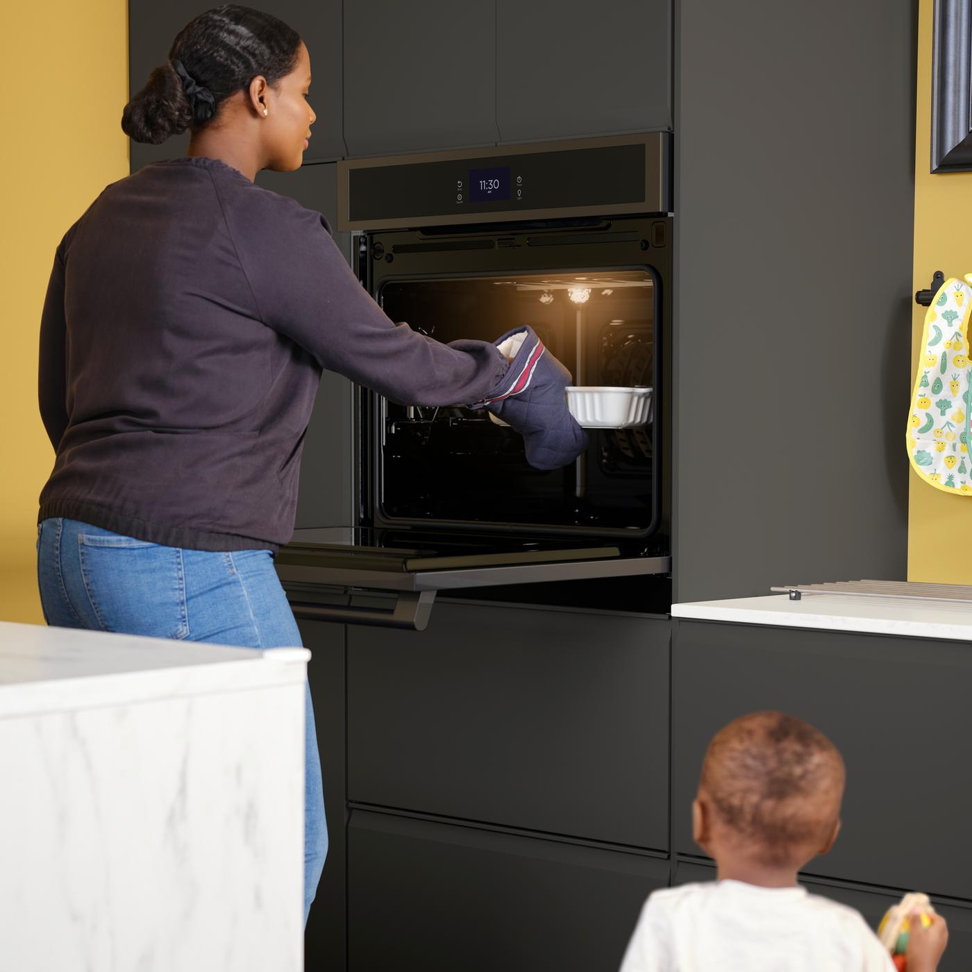 A toddler looks on as a woman takes an oven dish out of a HJÄLTEBY wall oven in a kitchen with matt anthracite fronts.