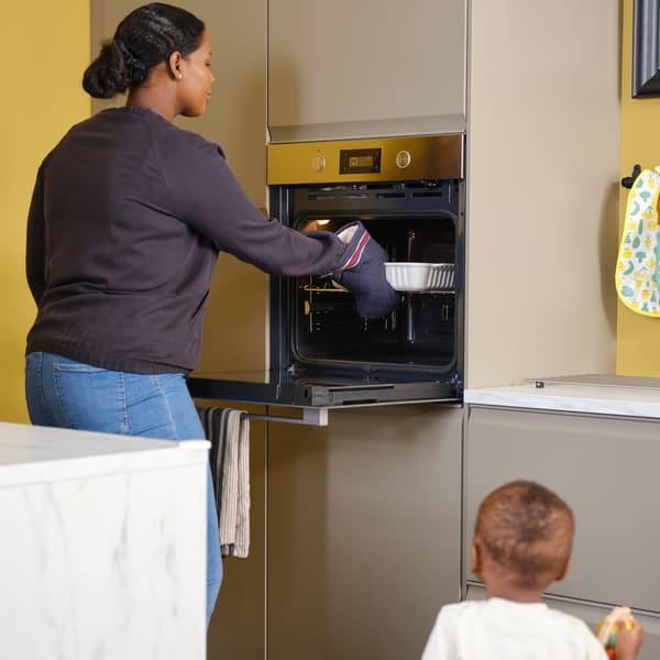 A toddler looks on as a woman takes an oven dish out of an ANRÄTTA forced air oven in a kitchen with matt dark beige fronts.