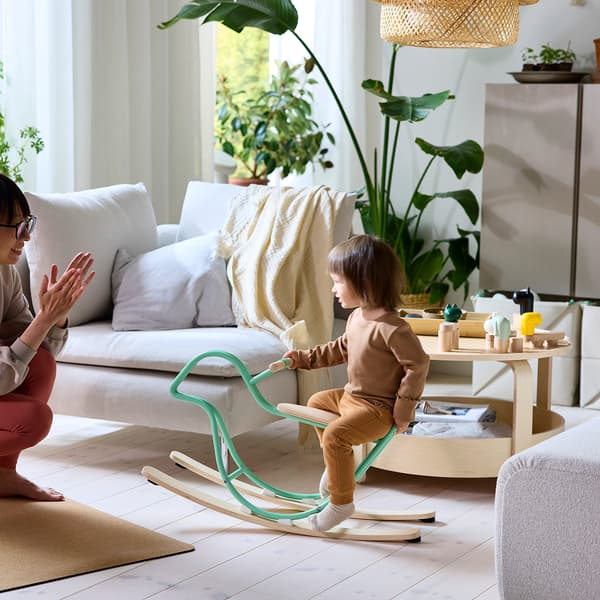 A toddler rides a rocking bird toy while her mother cheers her on.