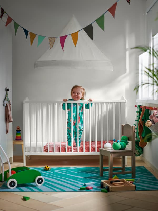 A toddler standing inside a white SMÅGÖRA crib, with kids' furniture and toys spread around the room.