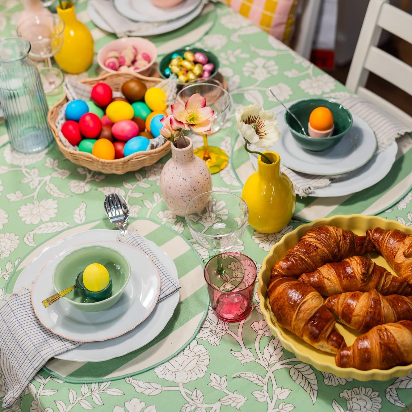 A top-view of an Easter table setting: colored eggs in a bowl, vases with flowers, and a cake.