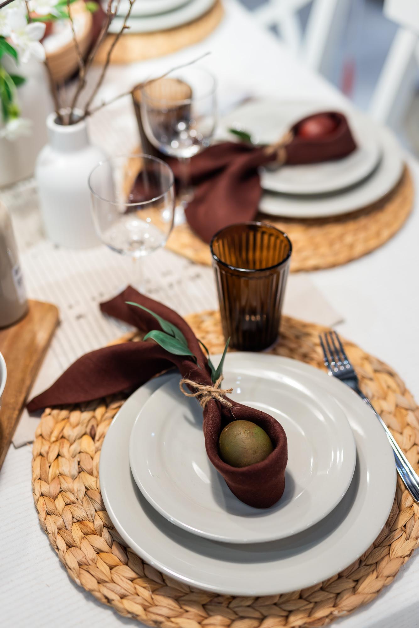 A top-view of an elegant Easter table setting: a woven place mat, white plates stacked on one another, and a coloured egg nestled in a decoratively folded napkin.