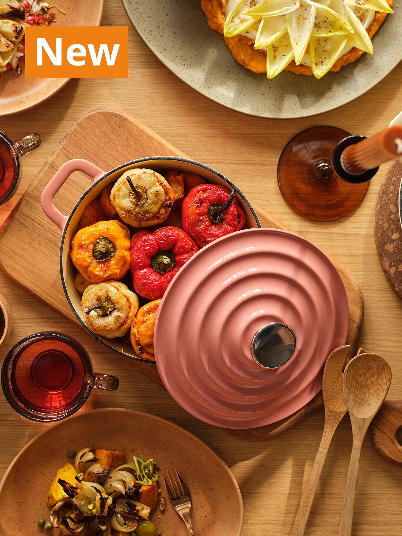 A top view shot of a table setting with VARDAGEN cast iron pot in light pink and a handle of GRÅRÄV tableware series in medium brown and medium green. Overlaying the image, orange tag that reads 'New' on the top left corner.