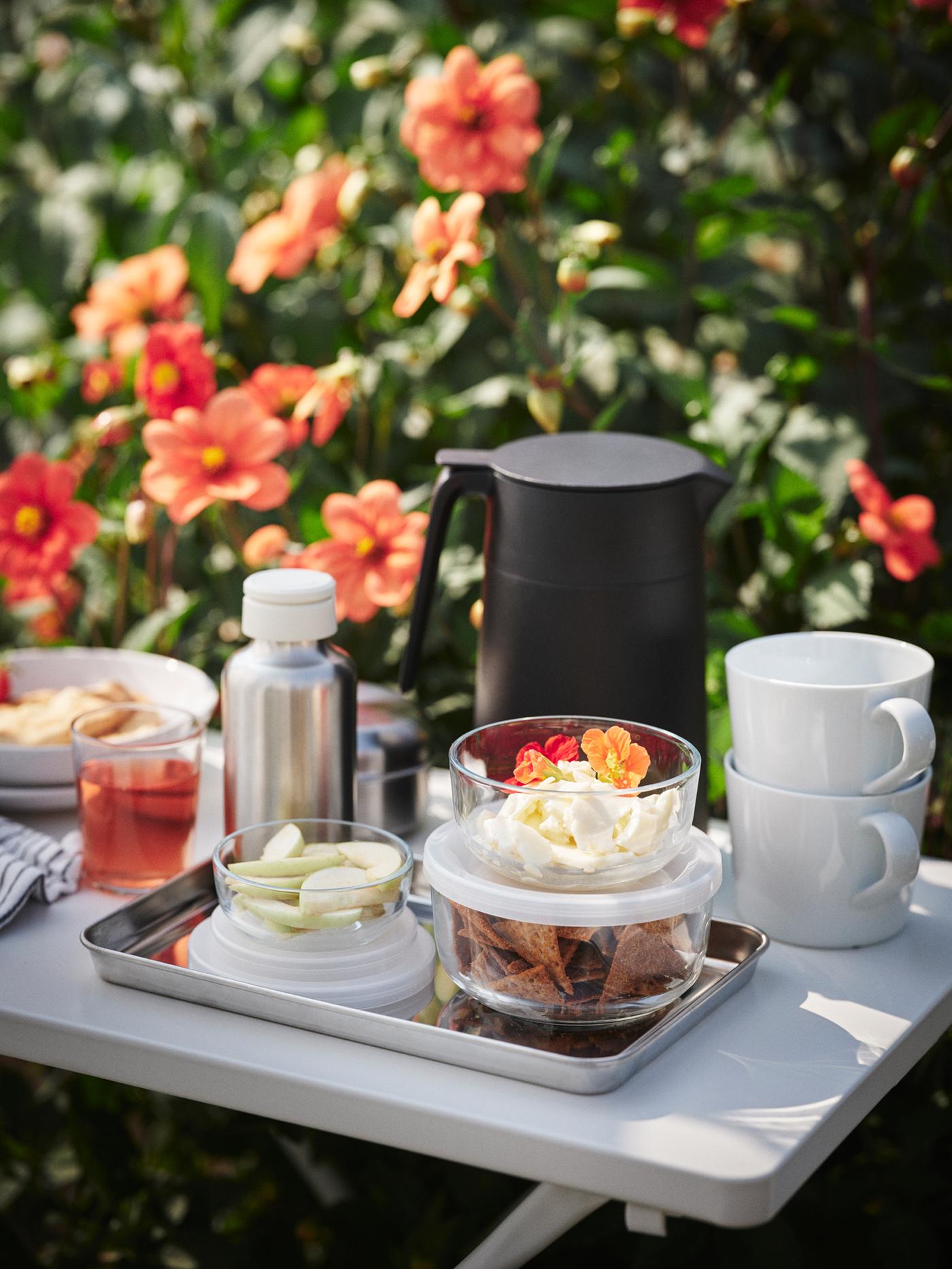 A TORPARÖ table is placed in a flowery garden, with snacks, mugs, a water bottle, and a vacuum flask on top.