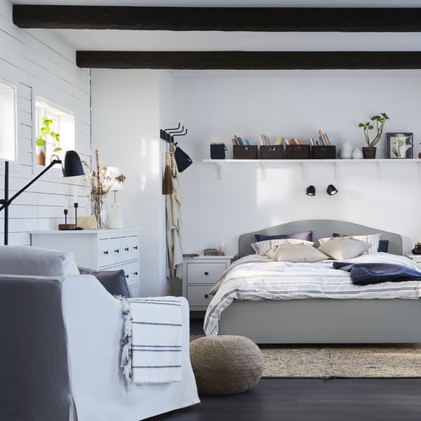 A traditional and coordinated bedroom with a beige HAUGA bed, a white armchair, white curtains and a dark blue-green desk.
