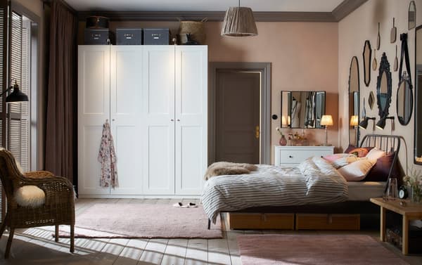 A traditional style bedroom with dusty pink walls and rugs, a gallery of mirrors above the bed, and two white wardrobes.