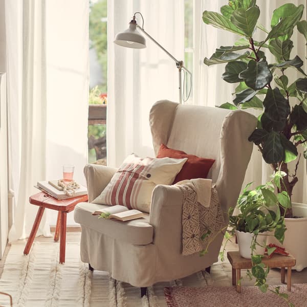 A traditional-styled STRANDMON wing chair with a natural linen slipcover has cushions and throws on it beside potted plants.