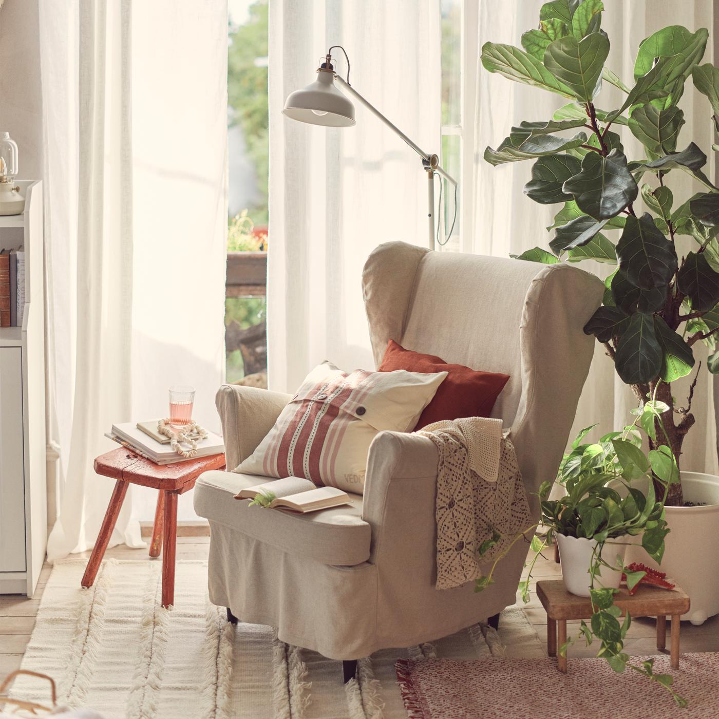 A traditional-styled STRANDMON wing chair with a natural linen slipcover has cushions and throws on it beside potted plants.