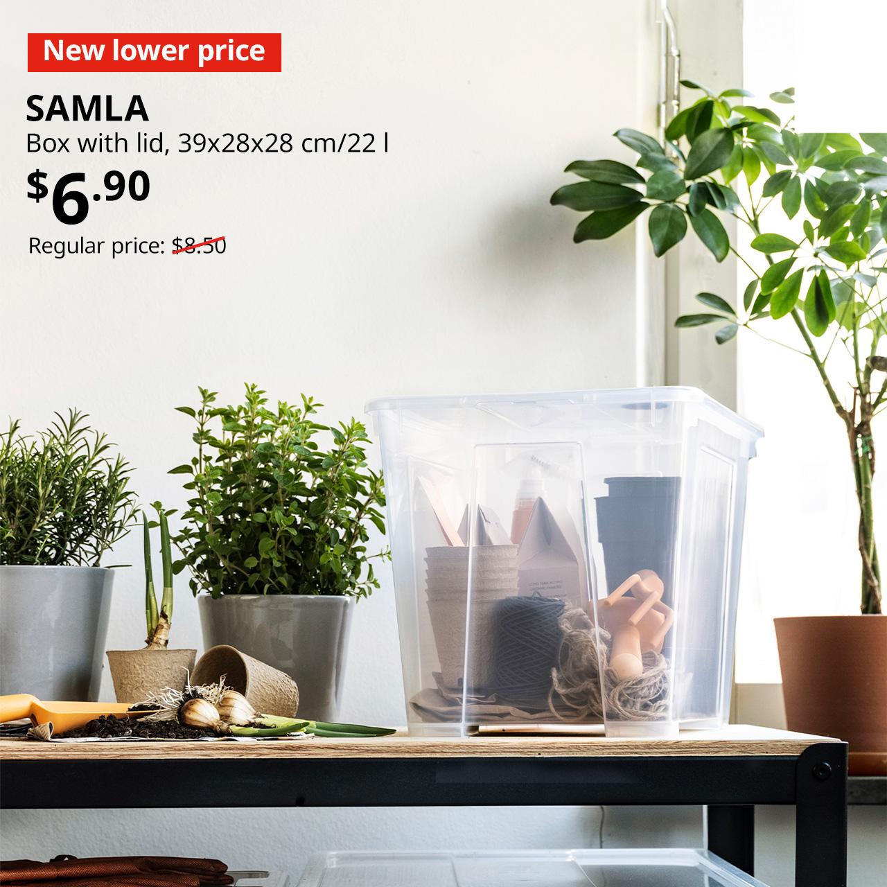A transparent SAMLA box with lid is placed on a black/pine plywood BROR work bench in the service yard alongside a pot of plant. In the top left corner, a red tag with white text reads 'New lower price'. Accompanying white text displays: 'SAMLA, Box with lid, 39x28x28 cm/22 l, $6.90, Regular price: $8.50'