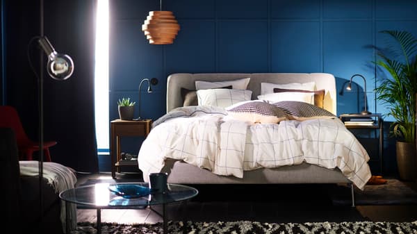 A TUFJORD upholstered bed in a blue bedroom covered with BREDVECKLARE bed linen. A DYKARKLOCKA pendant lampshade hangs above.