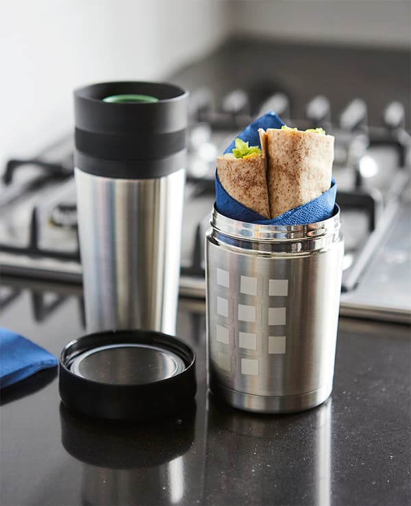 A vacuum flask thermos decorated with white stickers, filled with food, on a black worktop, near a cooker.