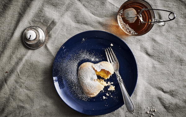 A vanilla heart biscuit and a fork on a dark blue plate, a mug with tea and a candle.