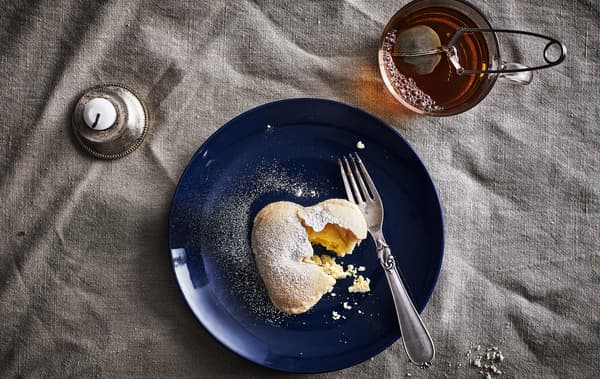 A vanilla heart-shaped biscuit and a fork on a dark blue dish, a cup of tea and a candle.