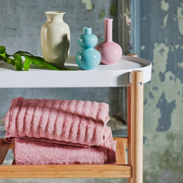 A variety of colored vases placed on top of a white and wooden side table. Light pink towels are placed on the wooden rack.
