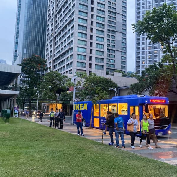 A vibrant blue IKEA bus is parked by a modern urban setting, with people walking nearby and high-rise buildings in the background.