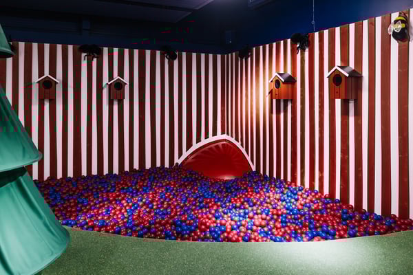 A vibrant indoor ball pit filled with red and blue balls, set against red-and-white striped walls decorated with birdhouses. A tunnel slide emerges from the corner, adding to the playful forest-themed design.