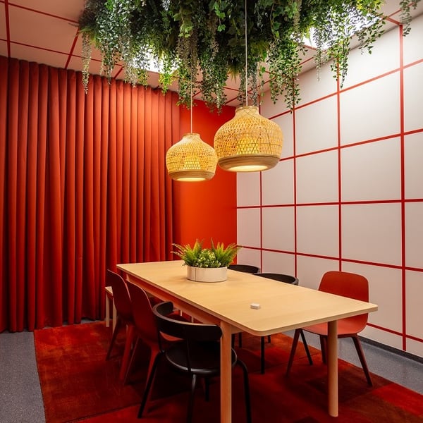 A vibrant meeting room features a rectangular light wood table surrounded by red and black chairs on a deep red carpet. Two woven pendant lamps hang above the table, suspended from a ceiling densely covered in cascading green plants. The left wall is covered with bold red curtains, while the right wall showcases a white grid pattern with red lines, creating a striking geometric contrast. A round planter with green foliage decorates the center of the table, enhancing the room's modern and nature-inspired aesthetic.