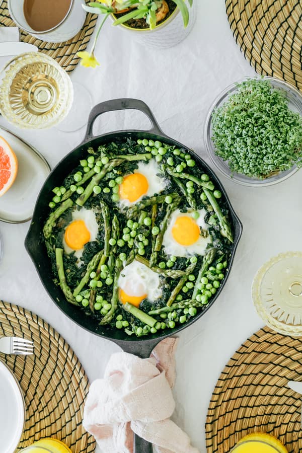 A vibrant skillet meal featuring asparagus, peas, and eggs on a white table with woven placemats, surrounded by dishes and green salad, evokes a fresh, cozy brunch.