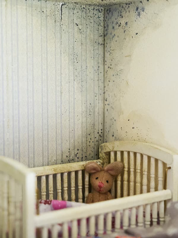 A view of the corner of a kids bedroom where a crib is standing right below a large mould stain on the wall and ceiling.