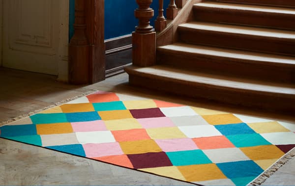 A VINDERÖD rug with a multicoloured, checkered pattern lies at the base of a staircase in dark wood with elaborate details.