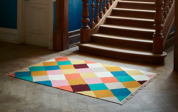 A VINDERÖD rug with a multicoloured, checkered pattern lies at the base of a staircase in dark wood with elaborate details.