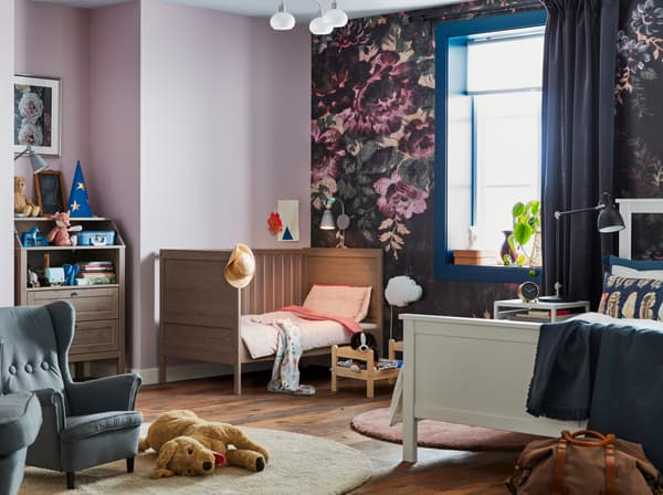 A violet and floral walled shared bedroom with parent and child, with IKEA SUNDVIK brown cot and changing table.