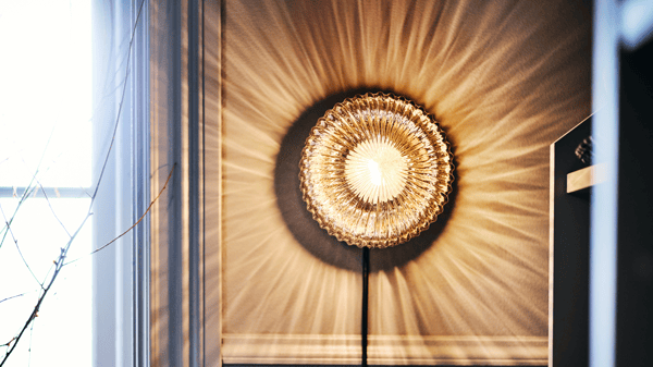 A wall-mounted lamp SOLKLINT with a ribbed glass shade, creating a stunning radial light pattern. The warm, golden light spreads out like sun rays, enhancing the room's ambiance. On the left, natural light filters through a nearby window, accompanied by thin branches. The overall scene is warm and inviting, combining natural and artificial light beautifully.
