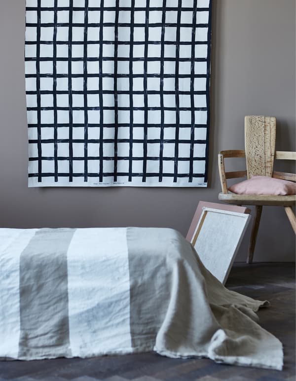 A wall textile with a grid pattern hanging on the wall behind a bed covered in a rough metre fabric painted with white stripes.