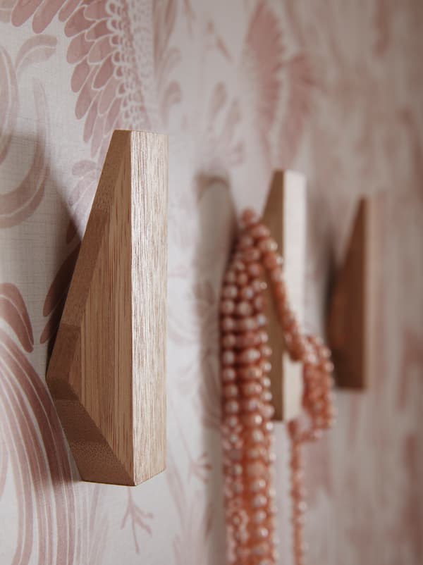 A wallpapered hallway wall with three SKUGGIS geometrically shaped bamboo hooks, and a beaded necklace hanging from one hook.