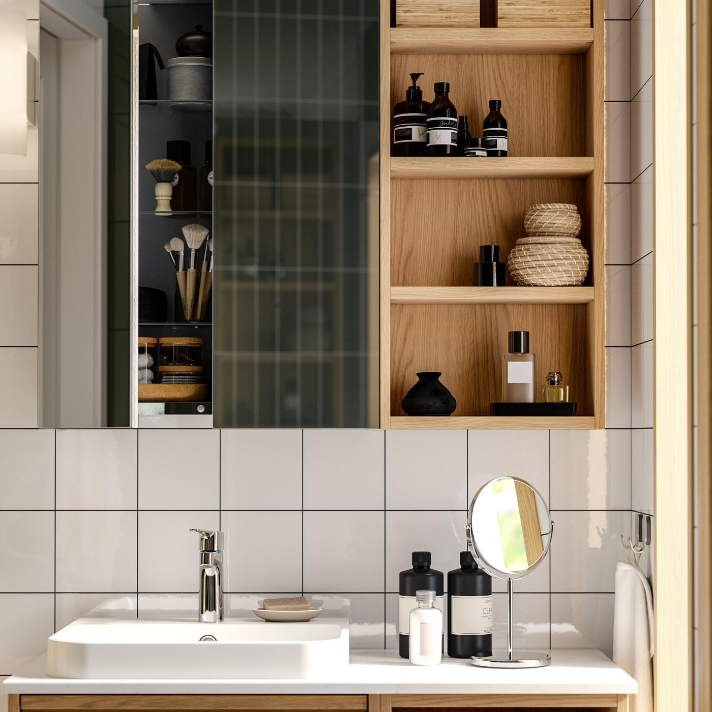 A washstand, mirrored wall cabinet with open shelving unit, baskets and jars, towels on hooks in a white-tiled bathroom.