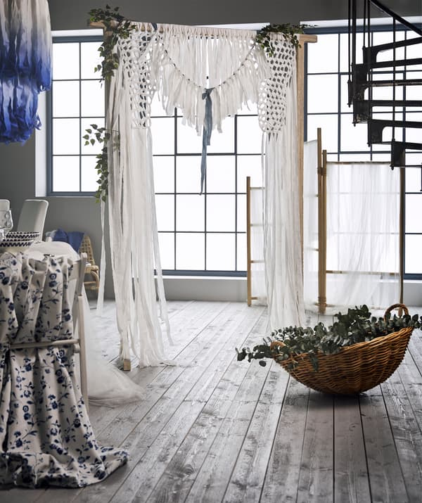 A wedding curtain backdrop made from white cotton fabric strips and greenery tied to a TÄNKVÄRD rattan room divider.