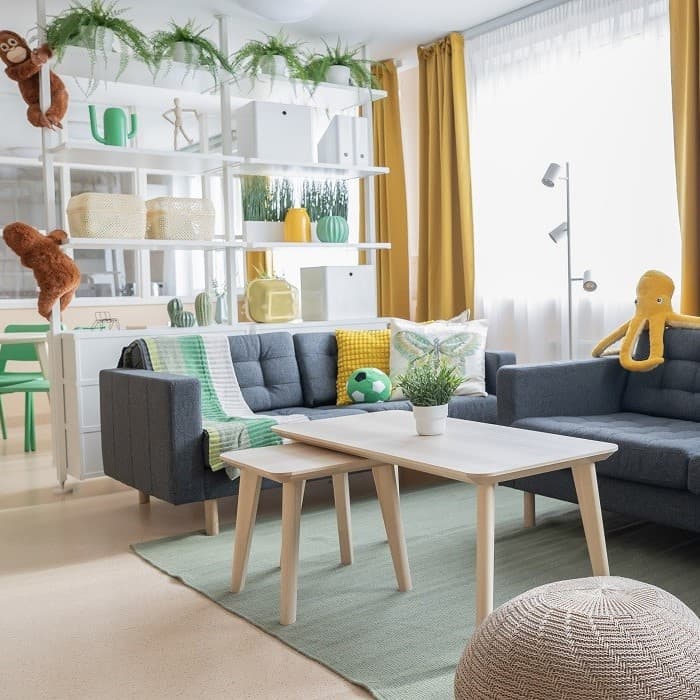 A welcoming shared lounge in a crisis centre with two sofas facing a coffee table, sunlight streaming through tall windows, and leafy plants lining open shelves.