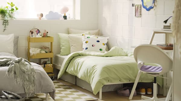 A well-lit, cozy dorm room with a single bed covered in a green duvet and polka dot pillows. A white chair, a small yellow side table with various items, and a shelf with toys and books are visible.